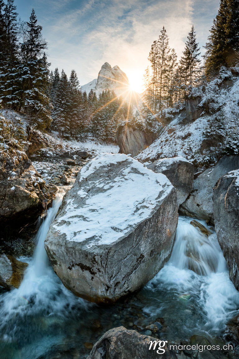 Winter picture Switzerland. Winter sun shining through the forest at Wetterhorn over Rychenbach creek in Rosenlaui Valley. Marcel Gross Photography